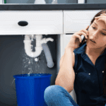 Homeowner on the phone making emergency plumbing call while water leaks under kitchen sink into a bucket.