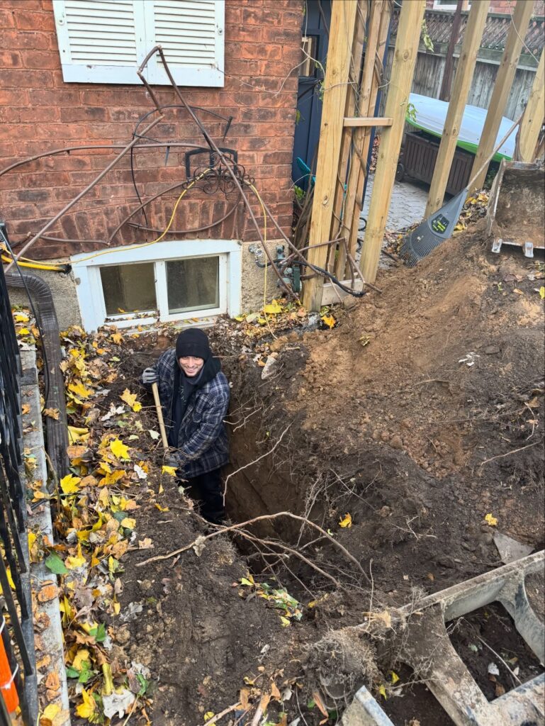 Technician standing in an excavated trench beside a house foundation during an underground pipe repair.