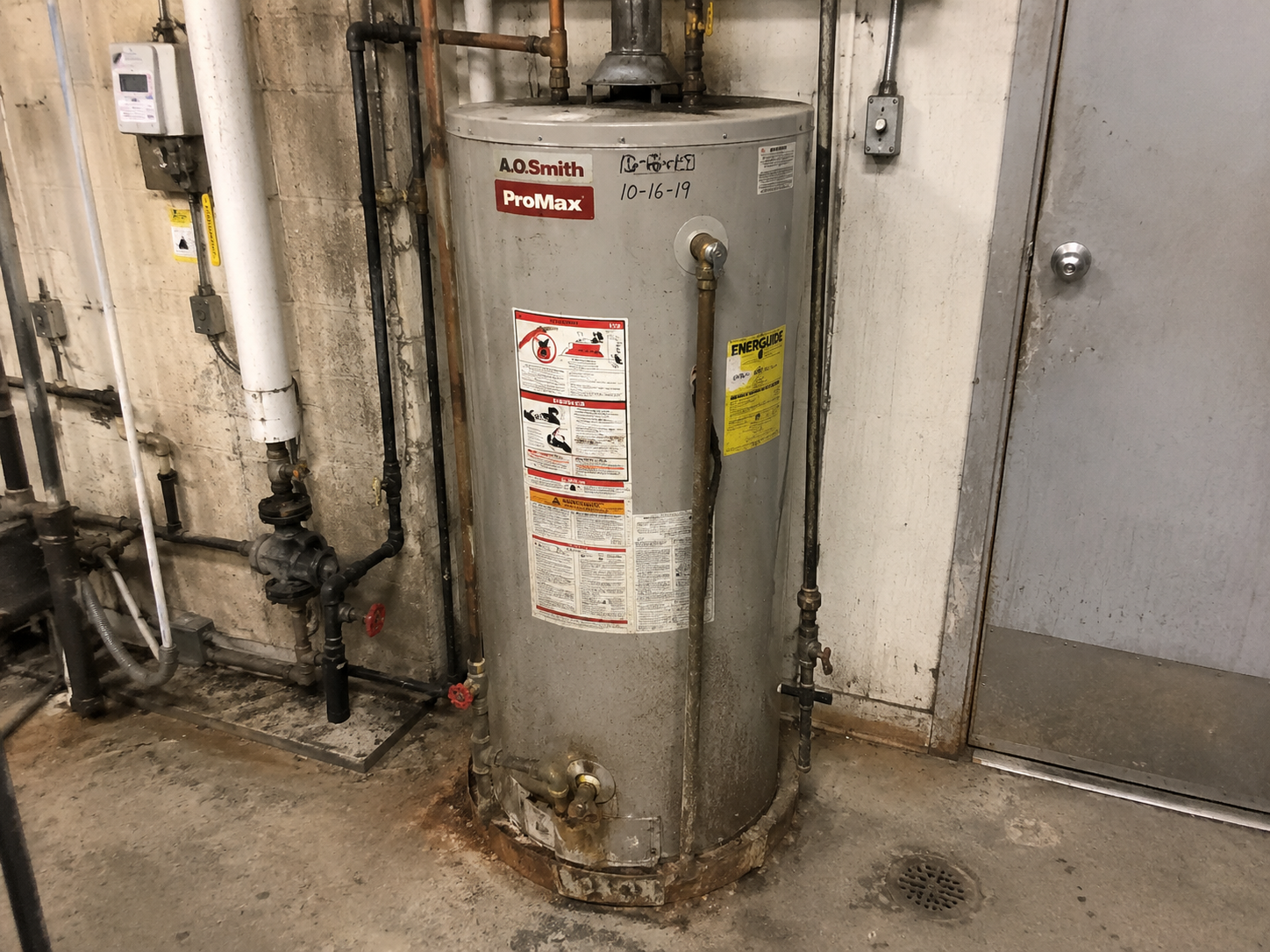 Older commercial water heater with rust and worn piping in a Hamilton utility room